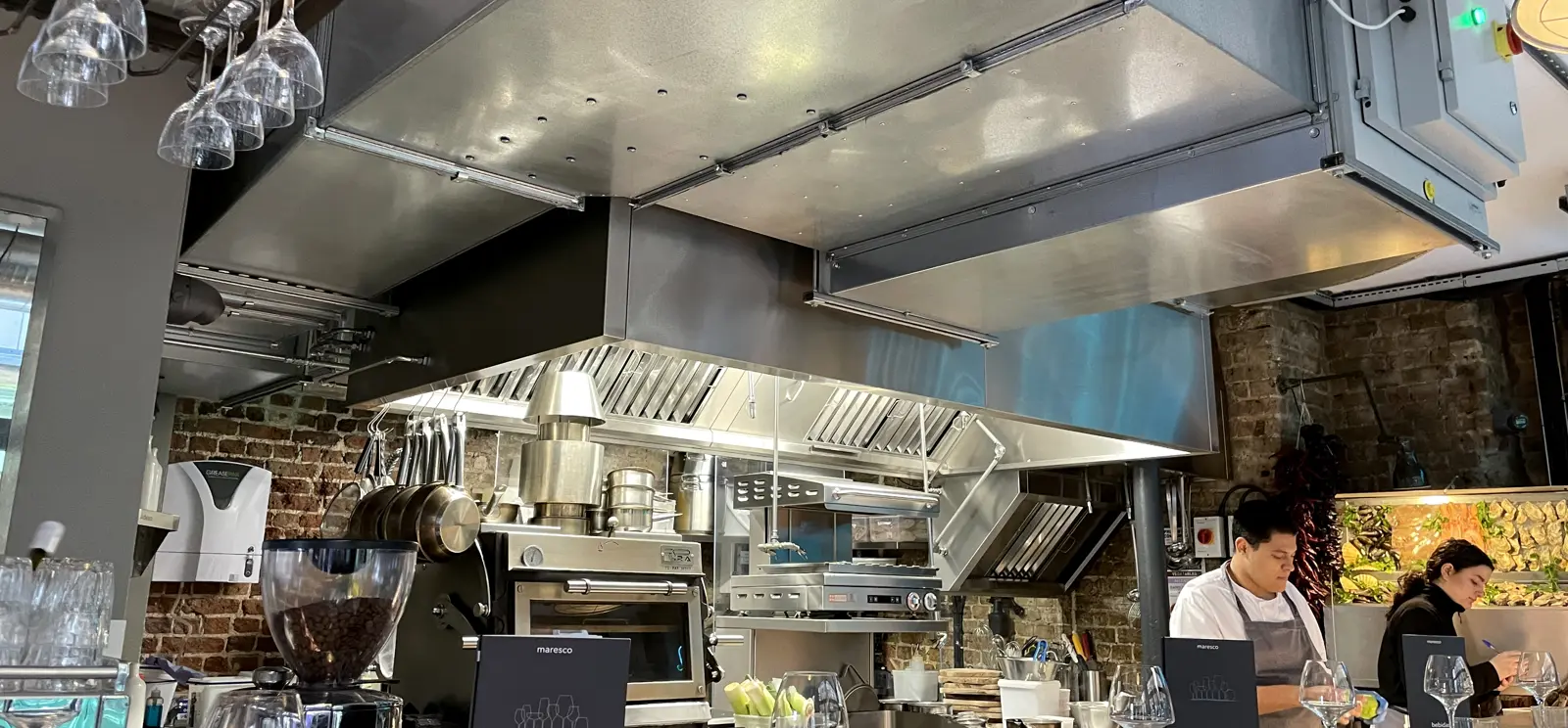 Front-of-house extract canopy with baffle filters and lighting above an open kitchen, with charcoal oven and grill below, at Maresco, Soho