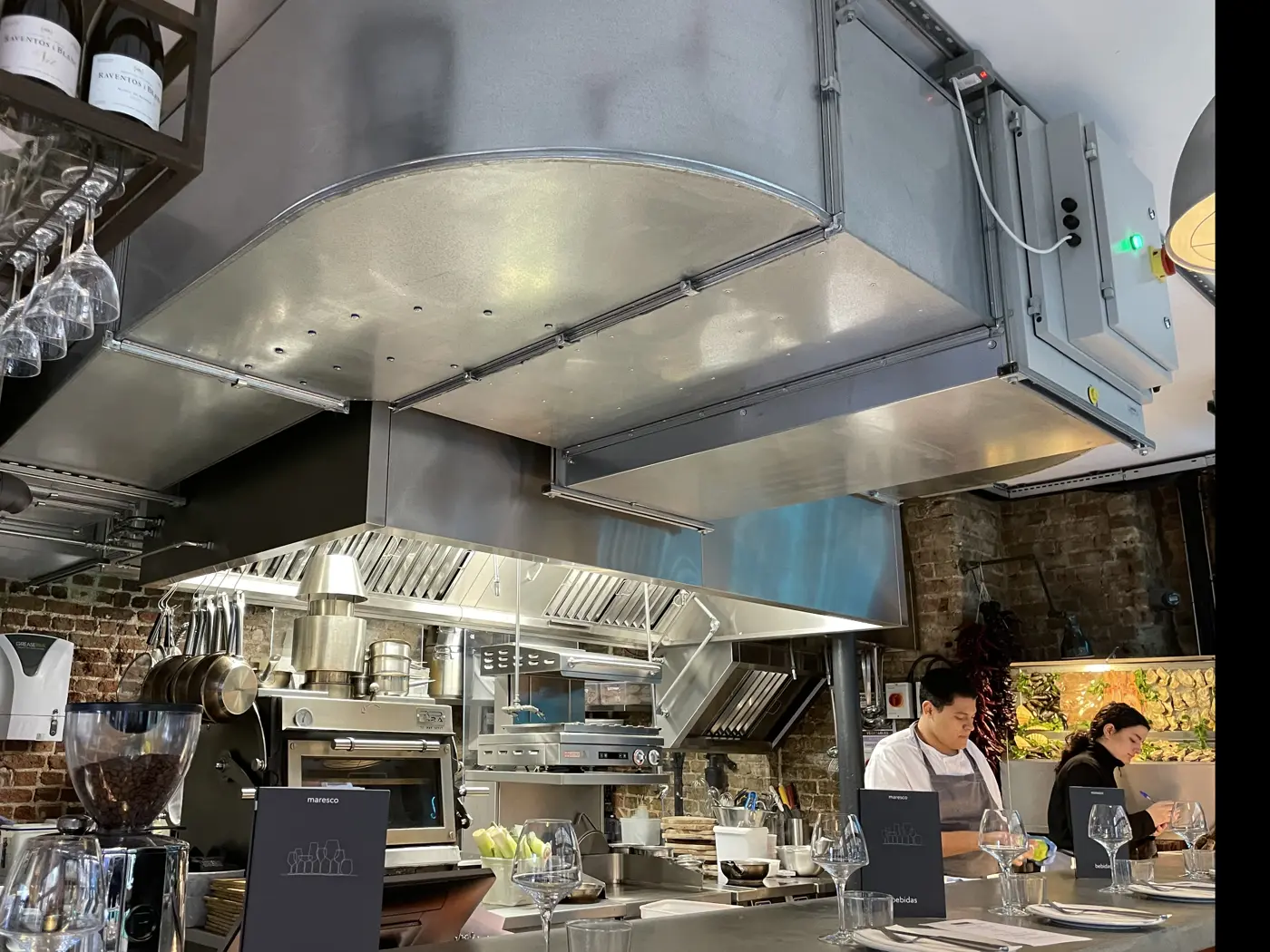 Front-of-house extract canopy with baffle filters and lighting above charcoal and grill cooking in an open kitchen